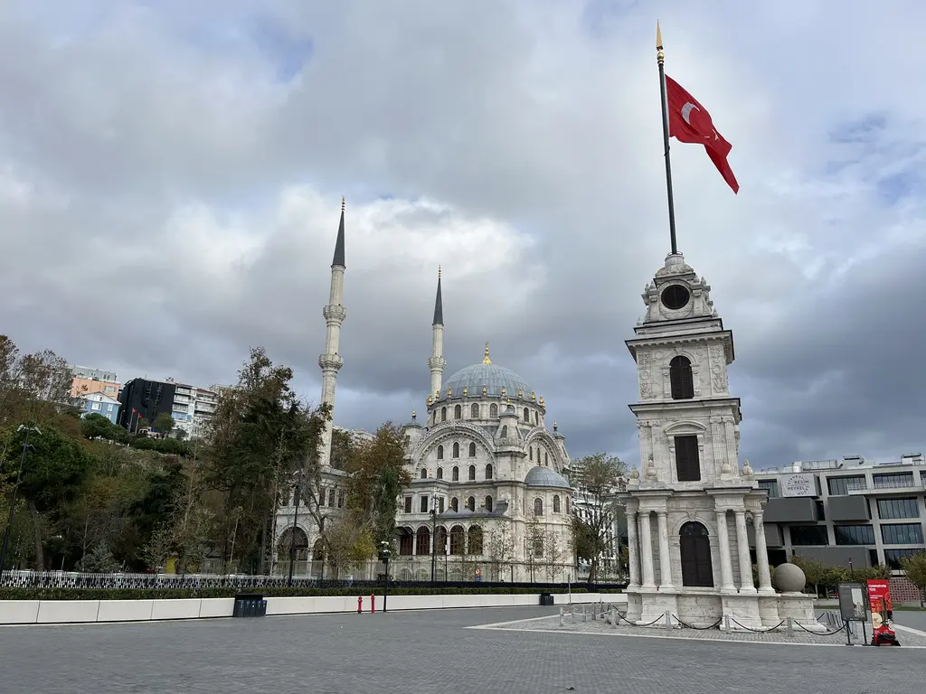 Tophane'de bulutlu bir günde meydan; sağda dalgalanan Türk bayrağı ve saat kulesi, solda kubbeli cami ve iki minare.