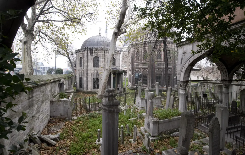 Şehzade Camii'nin haziresi; demir parmaklıkların ardında kavuklu mezar taşları, çınar ağaçları, arkada iki kubbeli türbe ve sağda revak.