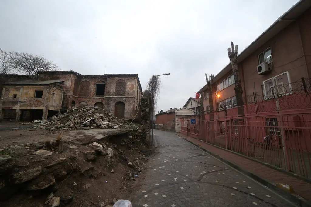 Balat'ta harap evlerin arasından geçen sokak; yıkık duvarlar, moloz, kırmızı tuğla bina ve kapalı gökyüzü.