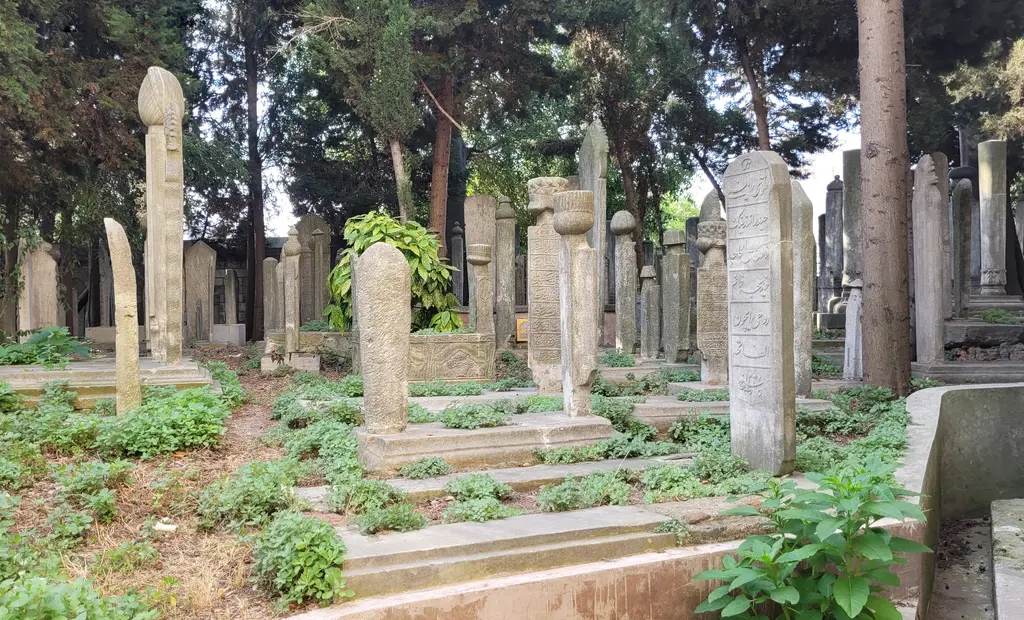 Külliyenin haziresi; ağaçların altında sıra sıra sarıklı mezar taşları, aralarında sütun gövdeleri ve arkada taş duvar.