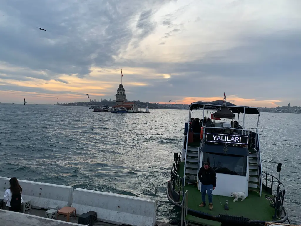 Salacak açığında Kız Kulesi; önde yanaşmış yeşil bir tekne, uzakta kule ve bulutlu akşam gökyüzü.
