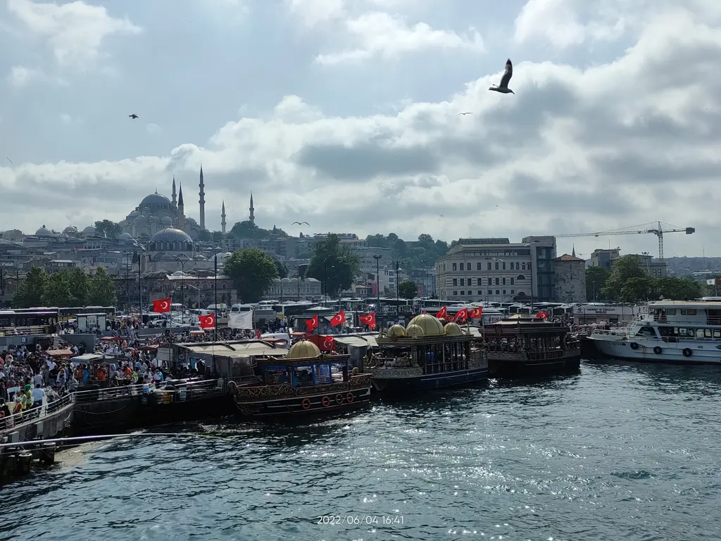 Eminönü kıyısında kubbeli süslü tekneler ve kalabalık; bayraklar, iskele, arkada Süleymaniye Camii ve bulutlu gökyüzü.