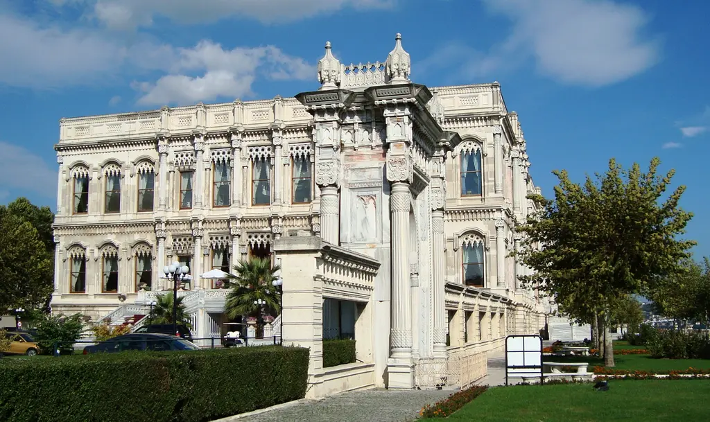 Çırağan Sarayı ve bahçesi; mermer saray cephesi, yanında oymalı taş kapı, önde çimenler ve ağaçlar.
