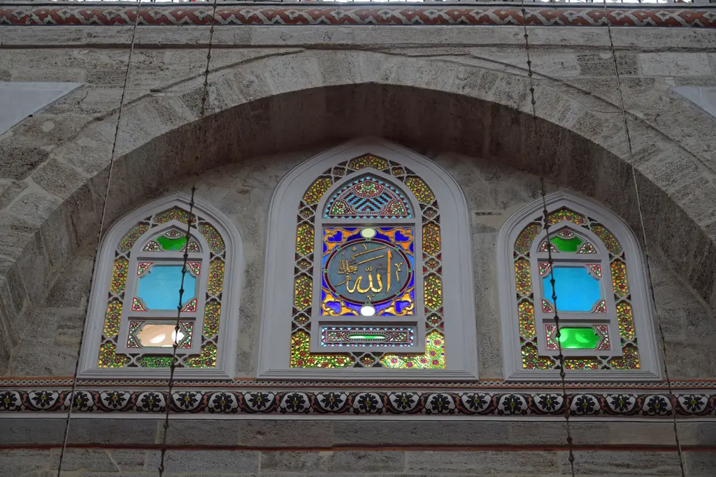 Mihrimah Sultan Camii'nde üç vitraylı pencere; alçı şebekeye oturtulmuş renkli camlar, ortadakinde yazı ve çevresinde badanalı duvar.