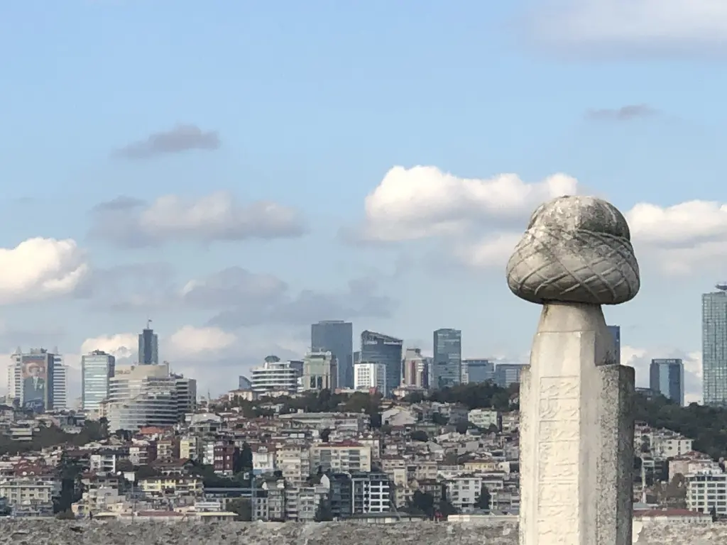 Şemsi Paşa Camii haziresinde sarıklı bir mezar taşı; arkada karşı kıyıda yükselen binalar ve bulutlar.