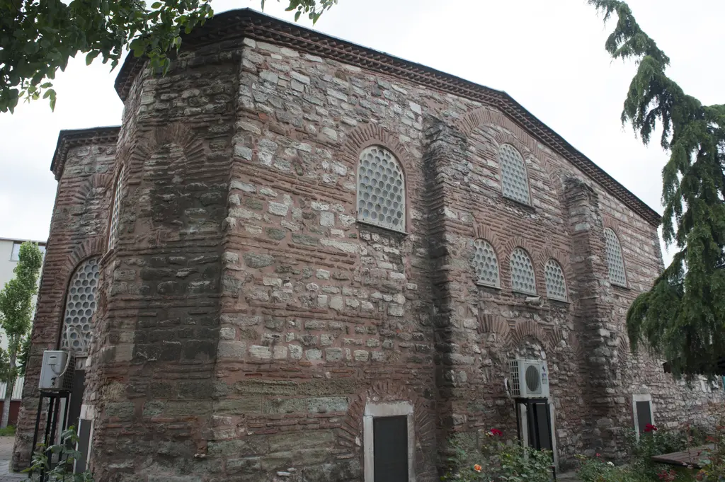 Atik Mustafa Paşa Camii'nin bahçeden görünüşü; sağda uzun yan duvar, solda çok yüzlü apsis çıkıntısı, kemerli şebekeli pencereler, önde gül fidanları ve çam ağacı.