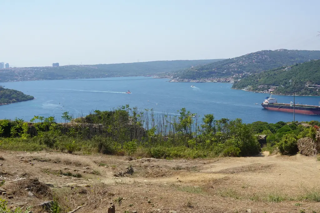 Yoros Kalesi'nden Boğaz; kuru otlu düzlüğün ardında lacivert deniz, geçen tanker ve iki kıyıda yeşil tepeler.