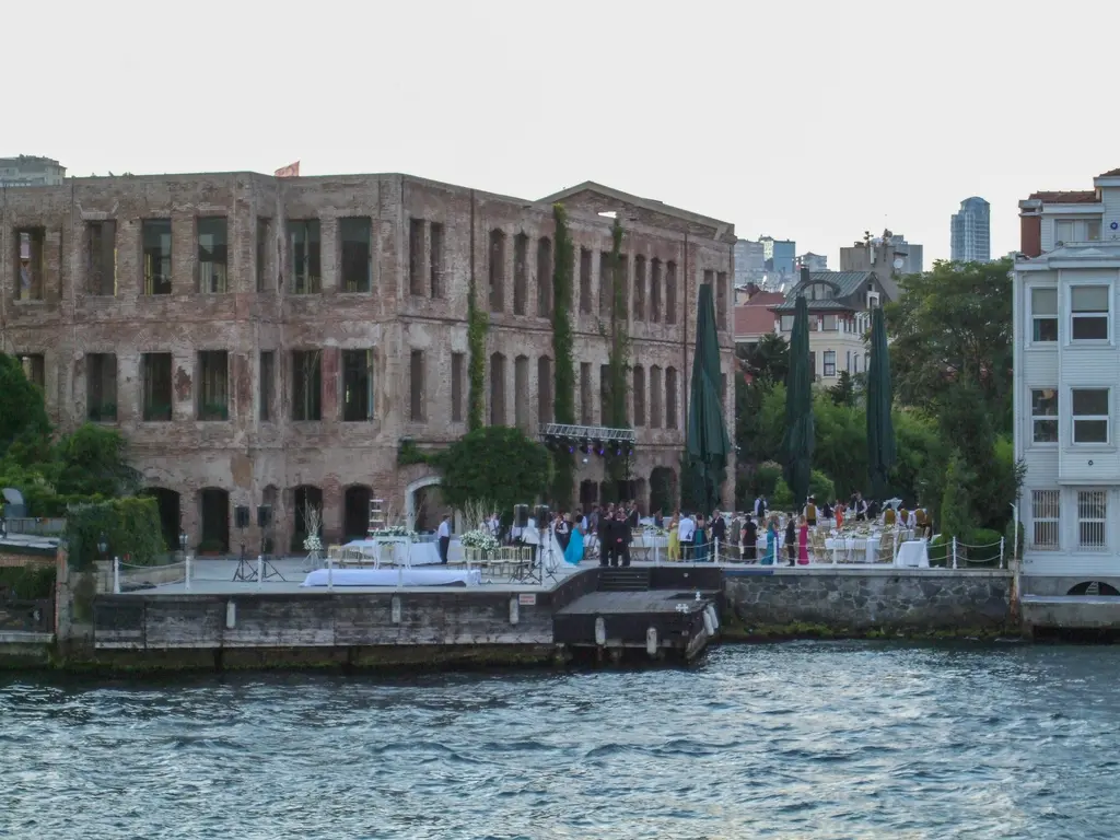 Boğaz'dan Ortaköy kıyısı; ortada yanık taş duvarlı Esma Sultan Yalısı, önünde bahçe ve kıyıda insanlar.