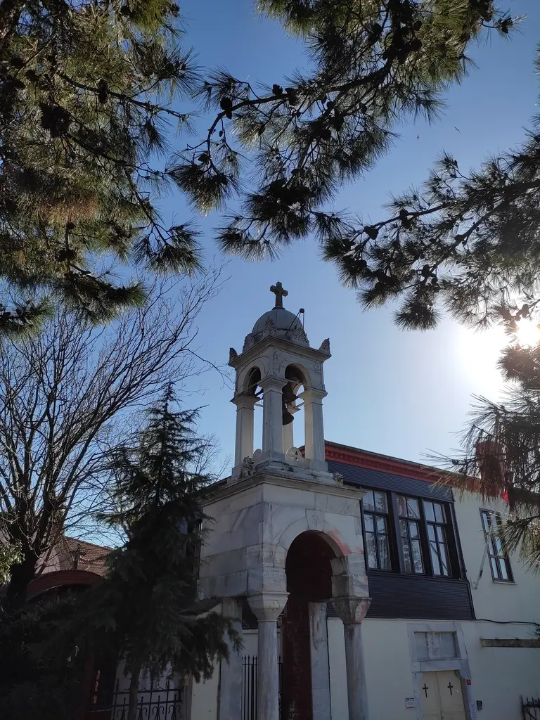 Aya Yorgi Manastırı; üstte çam dalları, ortada haçlı çan kulesi ve kemerli giriş, arkada güneş.