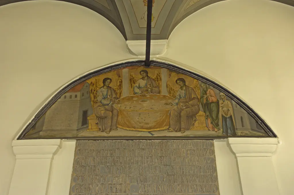 Kemer alınlığında altın zeminli resim; bir masanın çevresinde oturan üç kanatlı figür ve sağda ayakta duran iki kişi, altında Yunanca taş kitabe.