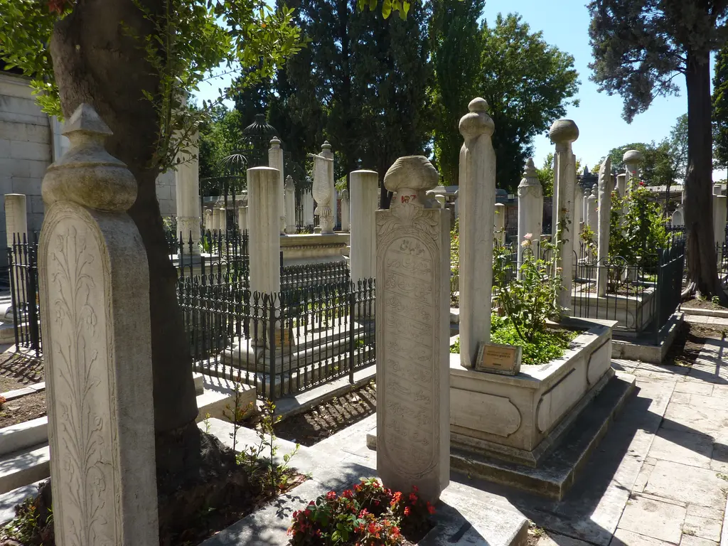 Fatih Camii'nin haziresi; sıra sıra beyaz mermer mezar taşları, tepelerinde oyma kavuklar, ferforje parmaklıklı lahitler, aralarında kırmızı güller ve serviler.