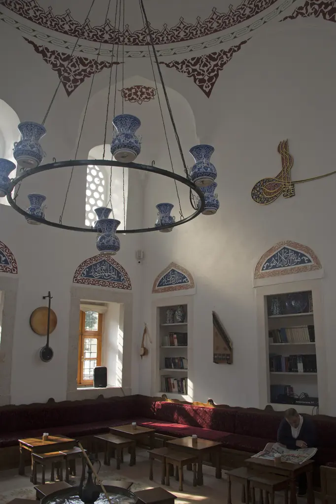 İç mekânın köşesi; beyaz duvarlarda kalem işi bordür, pencere altında kırmızı minderli sedir, yanda ahşap kitaplık ve rahleler.