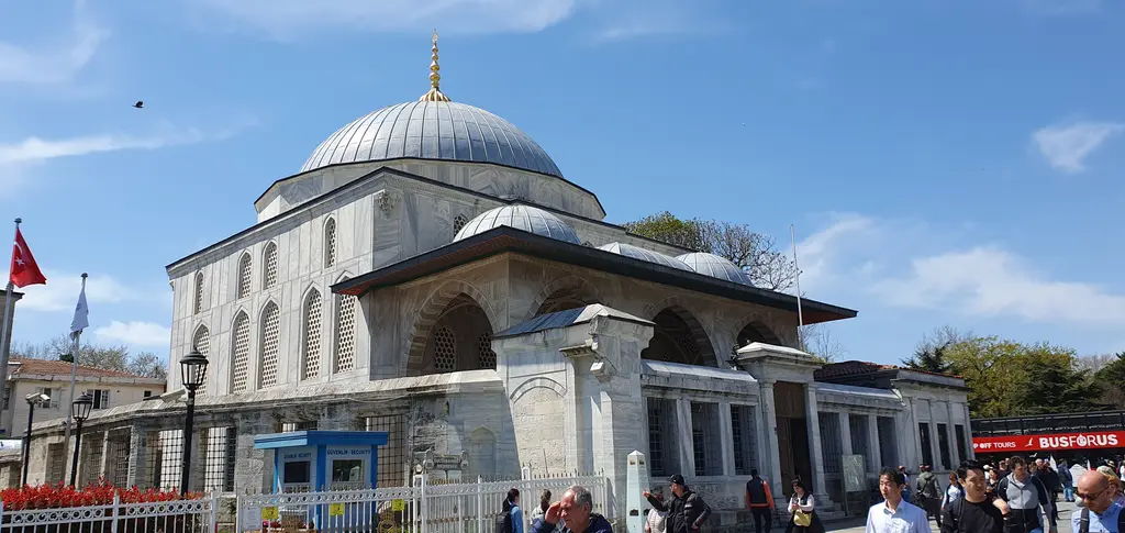 Sultanahmet Meydanı'ndan Sultan Ahmed Türbesi; kubbeli mermer yapı, avlu parmaklıkları, bayrak direkleri, ağaçlar ve önünden geçen kalabalık.