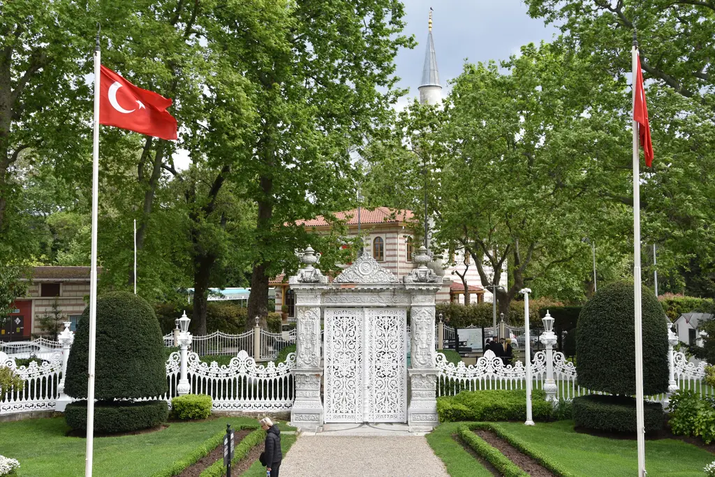 Küçüksu Kasrı'nın kara tarafındaki bahçe kapısı; iki bayrak direği arasında beyaz oymalı kapı, budanmış çalılar ve ağaçlar.