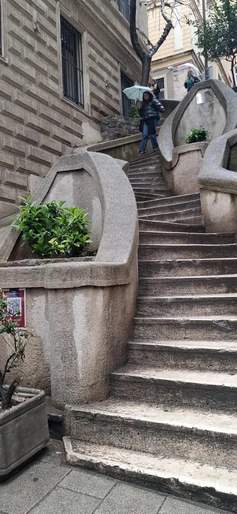 Kamondo Merdivenleri'nde yukarı çıkan insanlar; kıvrımlı basamaklar, saksıda bitki, iki yanda taş cepheli binalar.