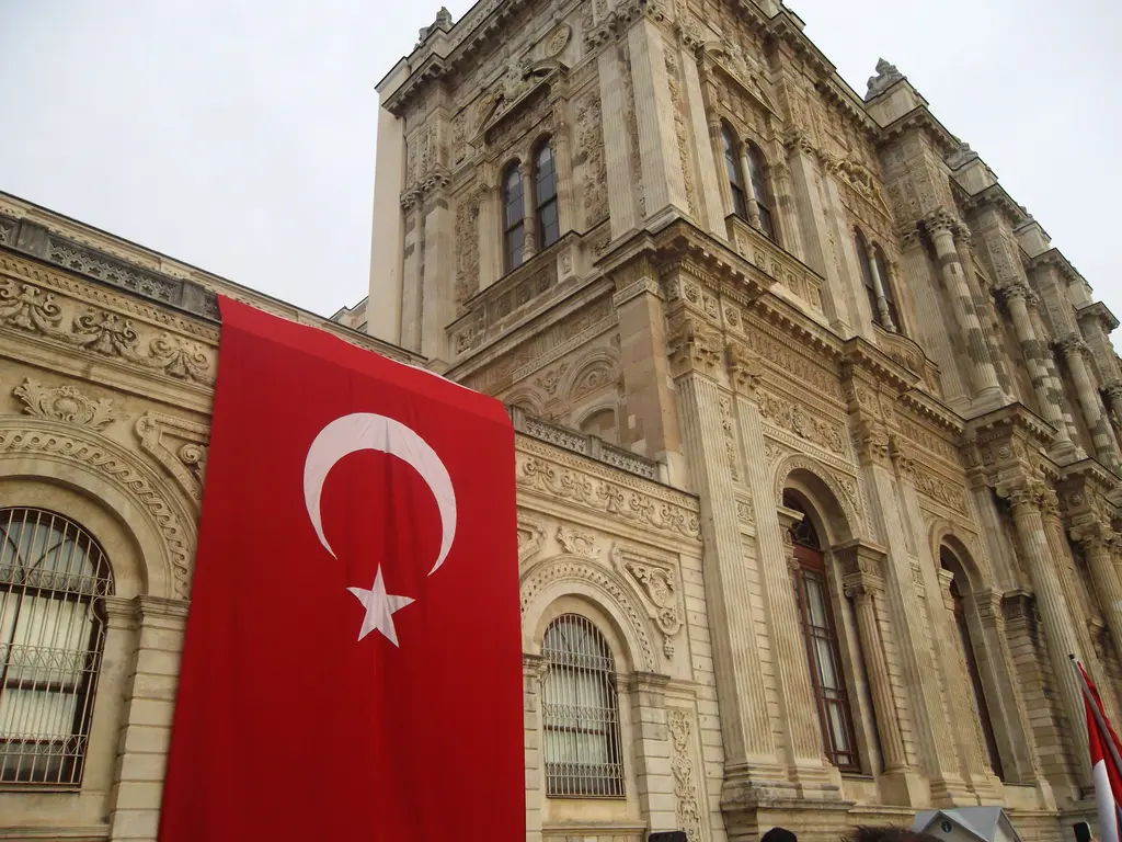 Dolmabahçe Sarayı'nın cephesi; oymalı taş süslemeler, kemerli pencereler, sütunlar ve önde asılı büyük Türk bayrağı.