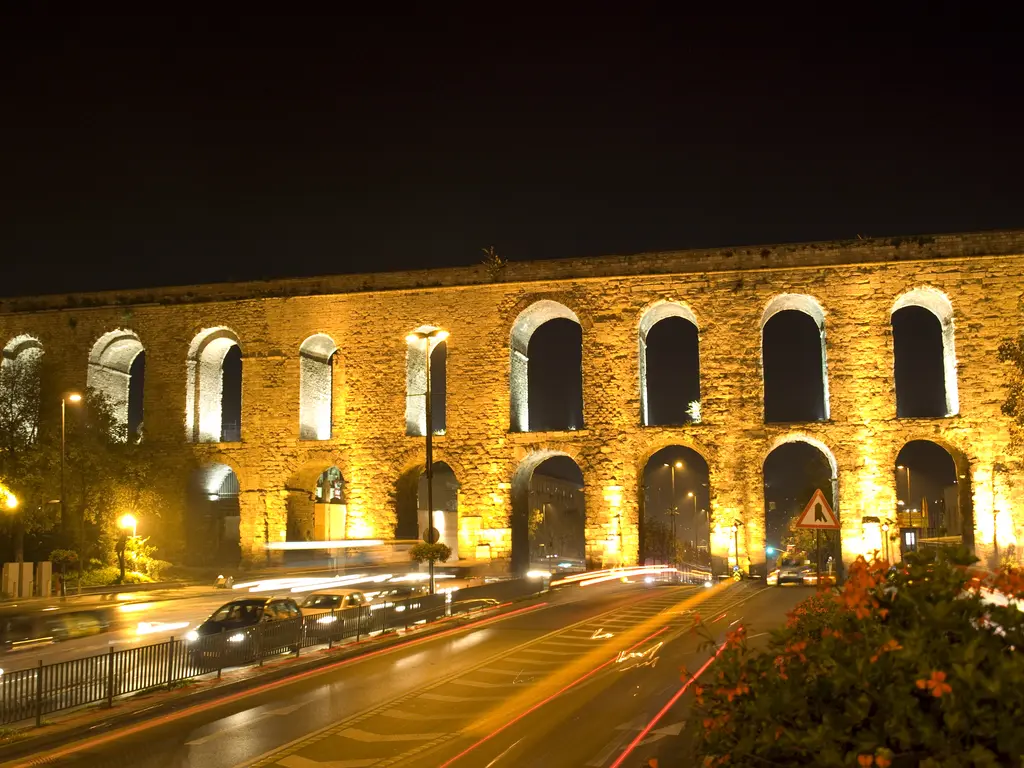 Bozdoğan Kemeri gece; dipten vuran projektörlerle altın sarısına boyanmış taş gövde, kapkara kemer gözleri, altta bulvarda kırmızı ve beyaz araç ışık izleri, siyah gökyüzü.