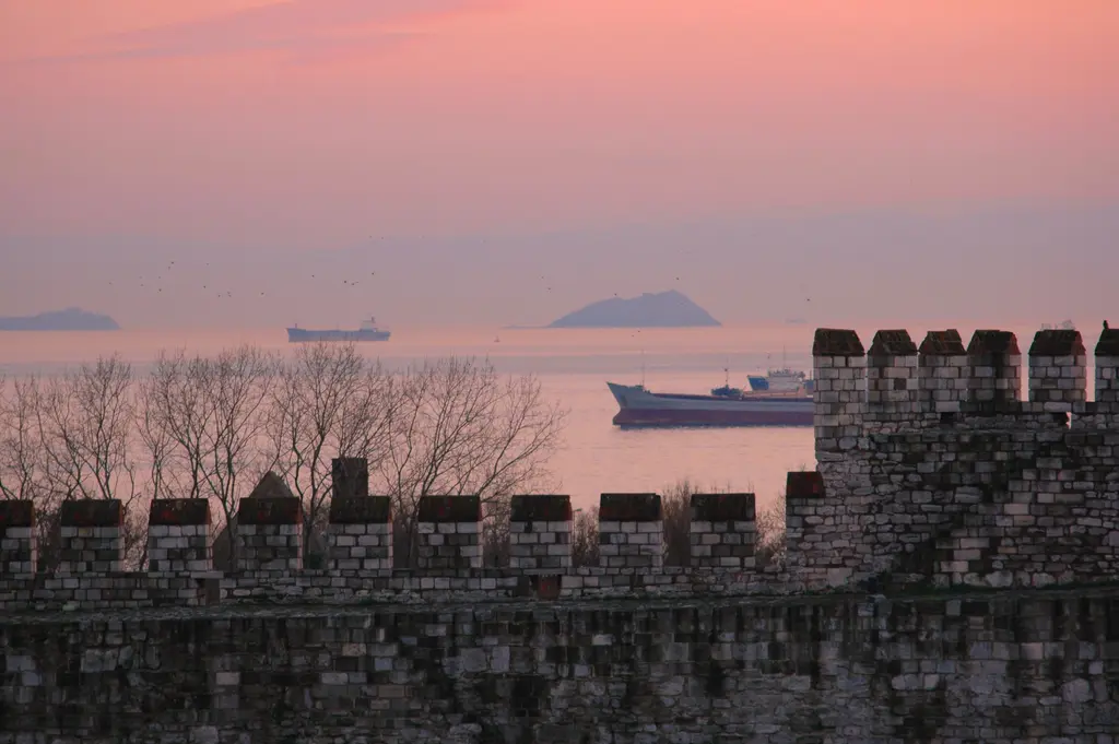 Yedikule Hisarı'ndan alacakaranlıkta Marmara Denizi; önde silüet hâlinde mazgallı sur korkuluğu ile yapraksız ağaçlar, pembe turuncu gökyüzü ve suda bir yük gemisi.