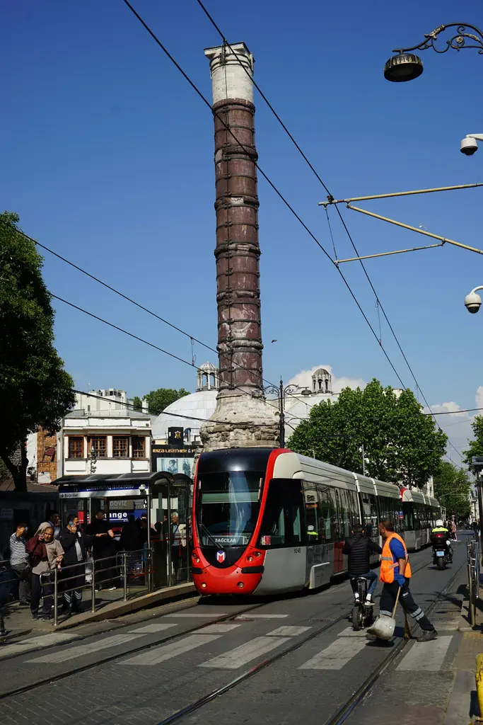 Çemberlitaş'ta kırmızı beyaz tramvay; sütunun önünden geçen araç, rayların üstündeki teller, arkada kaide ve ağaçlar.