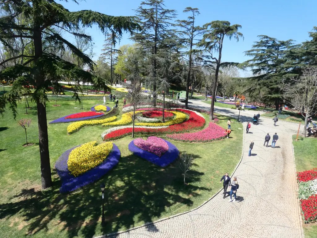 Emirgan Korusu'nda lale tarhı; çimenlikte kıvrılan sarı, kırmızı ve mor lale motifleri, sedirler ve yürüyüş yolunda insanlar.