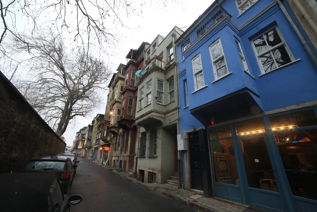 Balat'ta mavi boyalı ahşap ev; zemin katta ışıklı kafe, yanında gri ve krem evler, yokuş sokak ve çıplak ağaç.