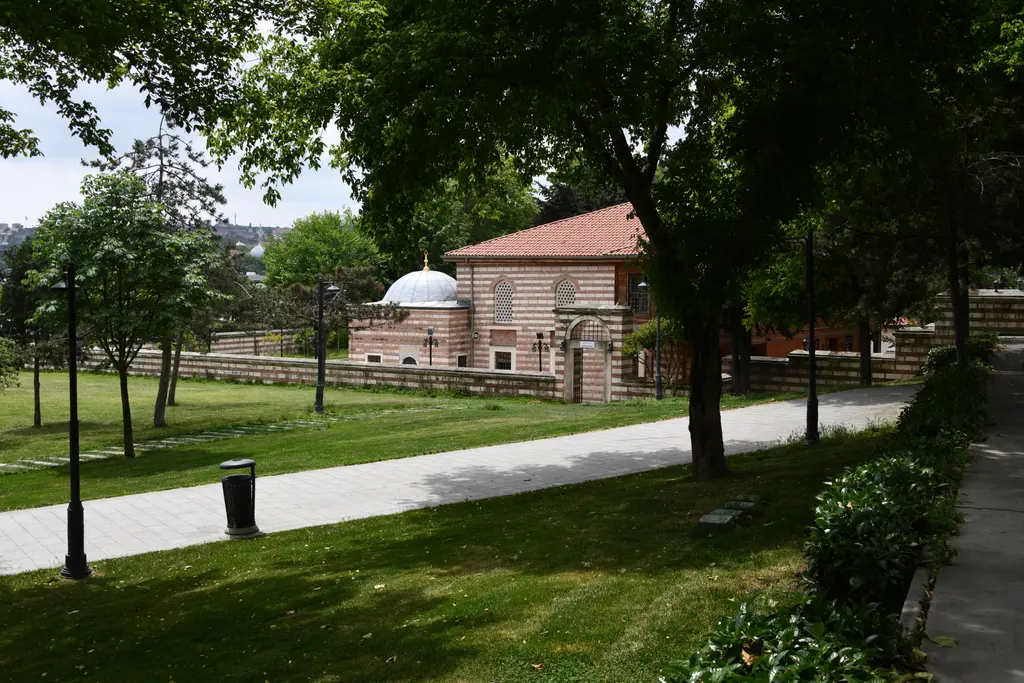 Şah Sultan Camii parktan; ağaçlar arasında kiremit çatılı cami, kubbeli türbe, çimenler ve parke yürüyüş yolu.
