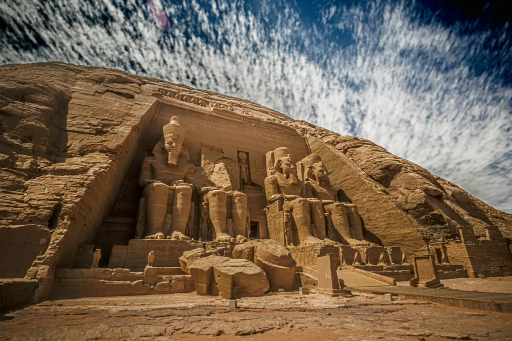 Mısır'da Abu Simbel tapınağının kaya cephesi; II. Ramses'in dev oturan heykelleri ve üzerinde ince bulutlu gökyüzü, alttan geniş açıyla.