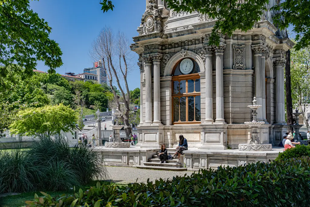 Dolmabahçe Saat Kulesi; süslü taş gövde, dört yüzünde saat kadranı, çevresinde bahçe, ağaçlar ve banklarda oturan insanlar.