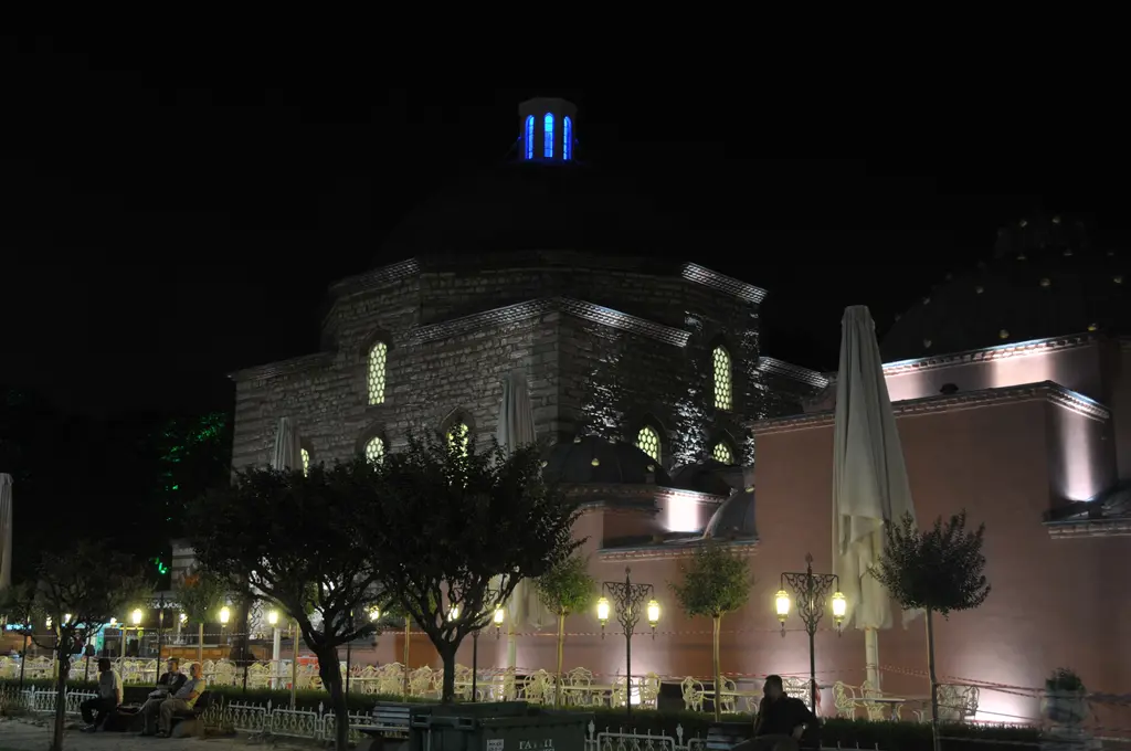 Gece aydınlatılmış hamam; sarı ışıkla yıkanan taş duvarlar, karanlıkta seçilen kubbeler ve önündeki boş yaya yolu.
