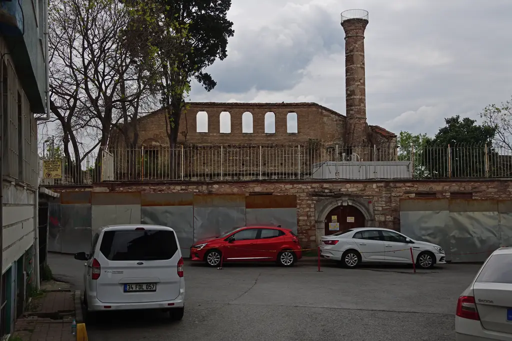 İmrahor İlyas Bey Camii restorasyon hâlinde; gri metal şantiye panoları, üstünde demir parmaklık, arkada kemerli pencereli duvar ile şerefeden yukarısı yıkık minare.