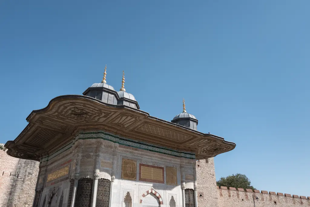 Çeşmenin Topkapı Sarayı'nın tuğla-taş örgülü sur duvarı önündeki görünüşü; geniş saçak, beş kubbecik ve mavi gökyüzü.