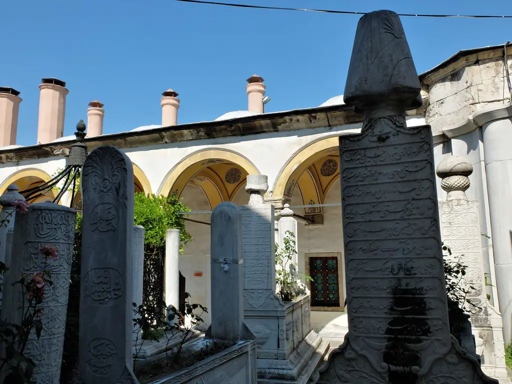 Mihrişah Valide Sultan İmareti'nin avlusu; kubbeli revaklar, yüksek bacalar, kurşun kubbeler, çiçekli bahçe ve taş döşeli yol.