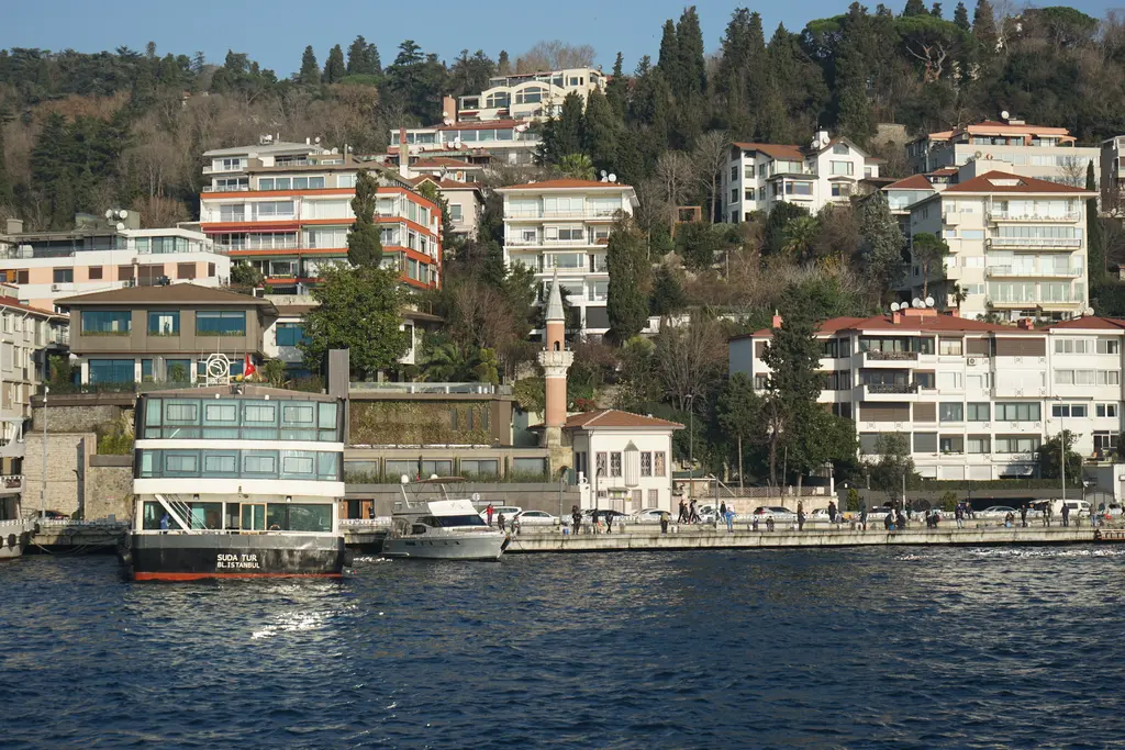 Bebek'te denizden Kayalar Mescidi; yamaçta ağaçlar ve evler arasında ince minare, önde rıhtım ve tekne.