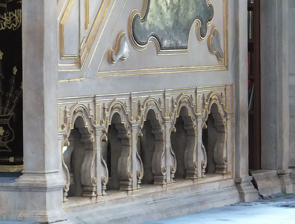 Nuruosmaniye Camii'nin minberi yandan; mermer korkulukta küçük kemer dizisi, kıvrımlı konsollar, üstte yaldızlı silme ve solda siyah zeminli hat panosu.