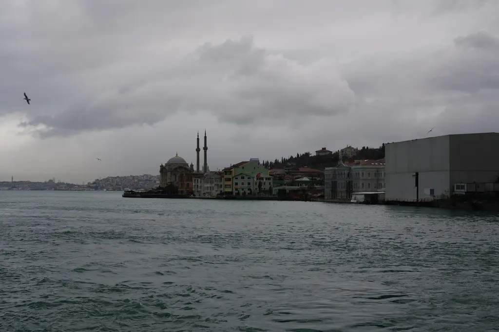Kapalı bir günde Boğaz'dan Ortaköy; dalgalı gri su, uzakta cami ve minareleri, kıyıdaki yapılar ve bulutlu gökyüzü.