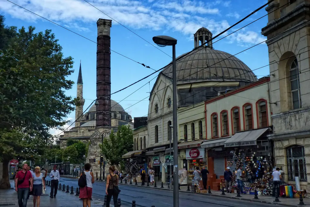 Divanyolu'nda Çemberlitaş sütunu; kararmış taş gövde, arkada bir minare, sağda hamamın kurşun kubbesi ve kalabalık kaldırım.