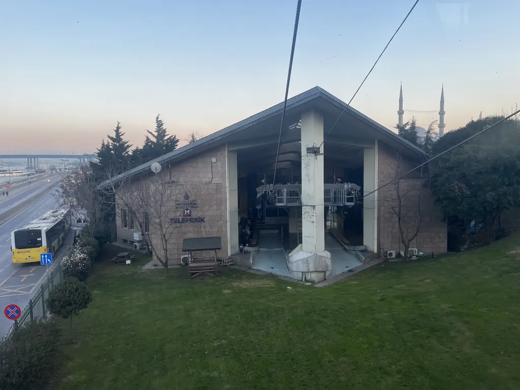 Eyüp teleferiğinin alt istasyonu; taş kaplı tek katlı yapı, kabin girişi, önünde çimen ve uzakta camiler.