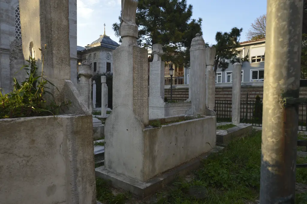 Ayazma Camii haziresinde mezarlar; taş sandukalar, uzun mezar taşları, yeşil bitkiler ve arkada ağaçlar.