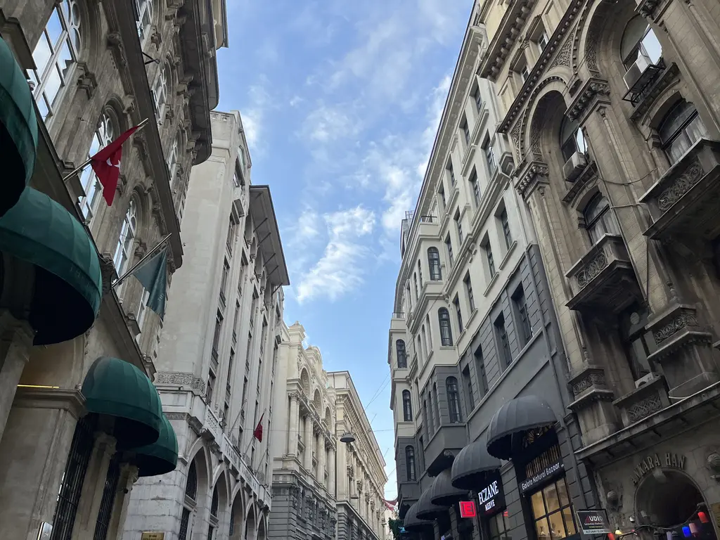 Bankalar Caddesi'nde hanlar; yeşil tenteli girişler, kabartmalı taş cepheler, asılı bayraklar ve mavi gökyüzü.