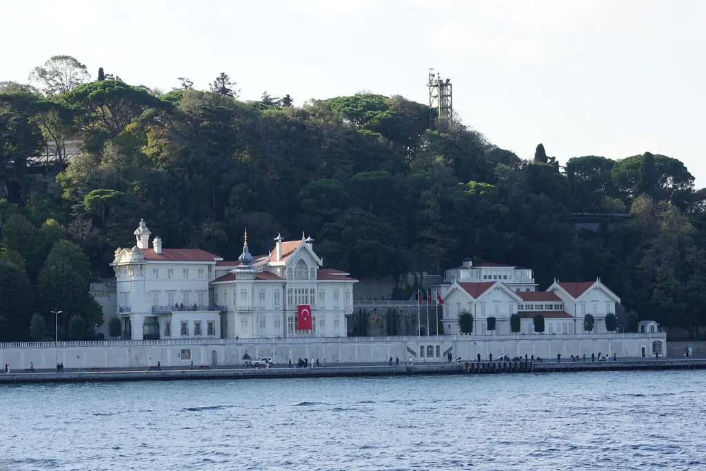 Boğaz'dan Huber Köşkü; kıyıda beyaz köşk ve yan binaları, arkada sırta tırmanan yoğun ağaçlık koru.