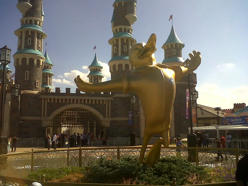 Vialand'de şato kapısının önünde iki kolunu açmış altın maskot heykeli; arkada turkuaz külahlı kuleler ve önde çevrili bir bahçe.