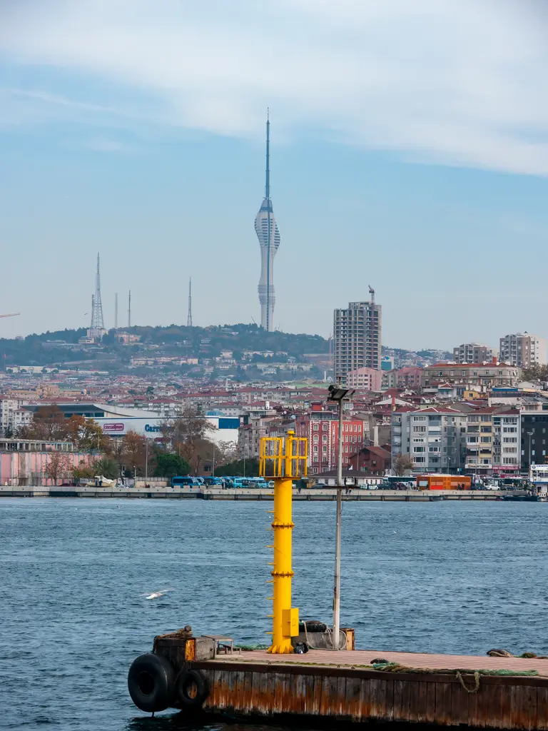 Kadıköy iskelesinden Çamlıca Kulesi; önde sarı şamandıra ve rıhtım, kıyıda binalar ve tepede kule.