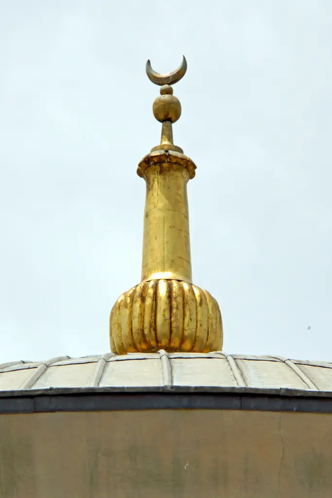 Kubbeciğin tepesindeki altın yaldızlı tunç alem; dilimli gövde, üstünde küre ve hilal, arkada açık gökyüzü.