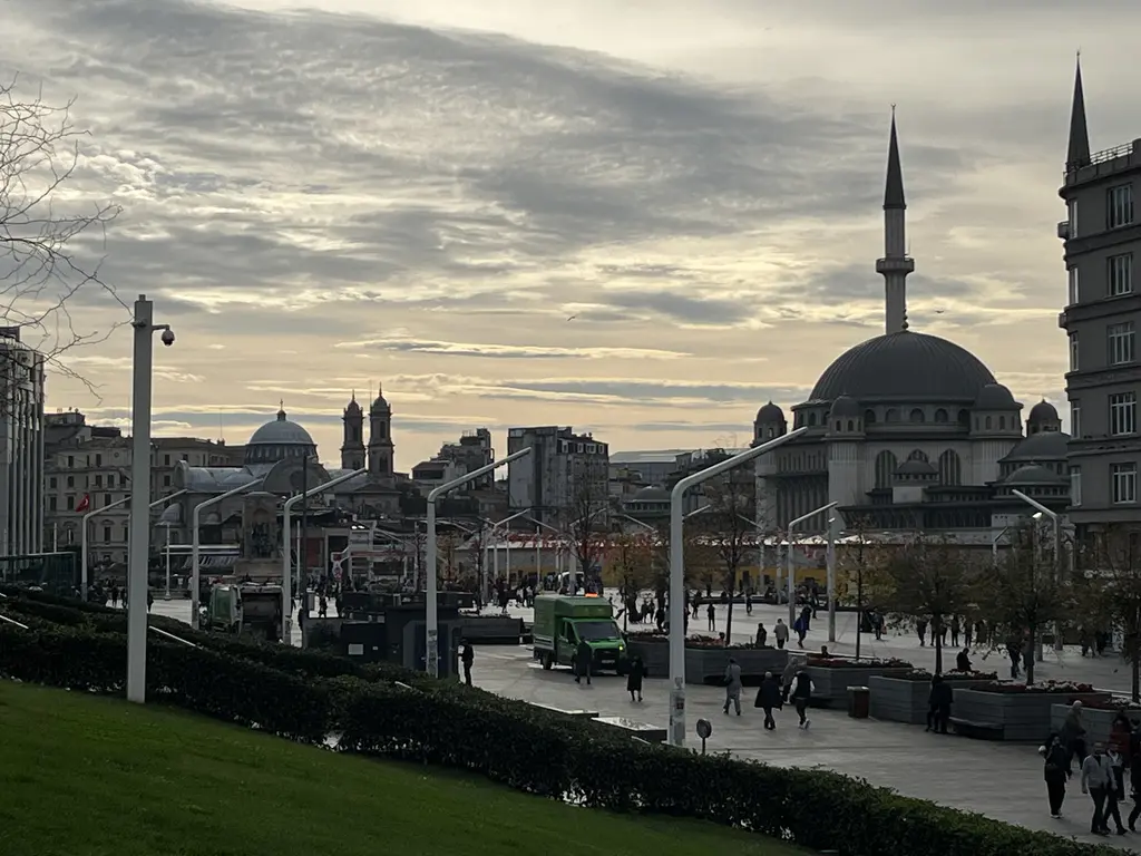 Gezi Parkı'nın kenarından Taksim Meydanı; akşamüstü bulutlu gökyüzü, sağda Taksim Camii, aşağıda yürüyen insanlar.