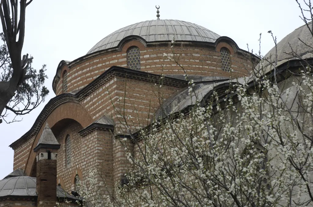 Rum Mehmed Paşa Camii'nin duvarları; taş ve tuğla sıralı örgü, büyük kemerler ve önünde beyaz çiçekli ağaç.