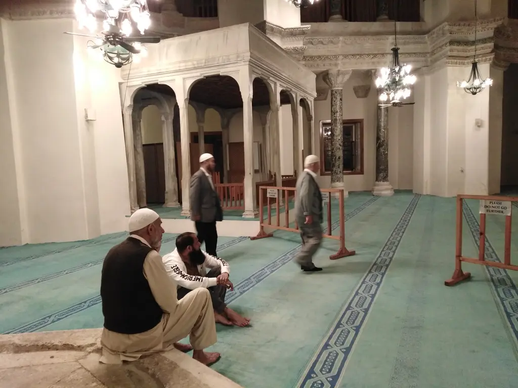 Küçük Ayasofya Camii'nde zemin kat sütun dizisi; deniz yeşili halı, önde taş sekide oturan üç kişi, arkada koyu damarlı mermer sütunlar, revak kemerleri ve asılı avizeler.