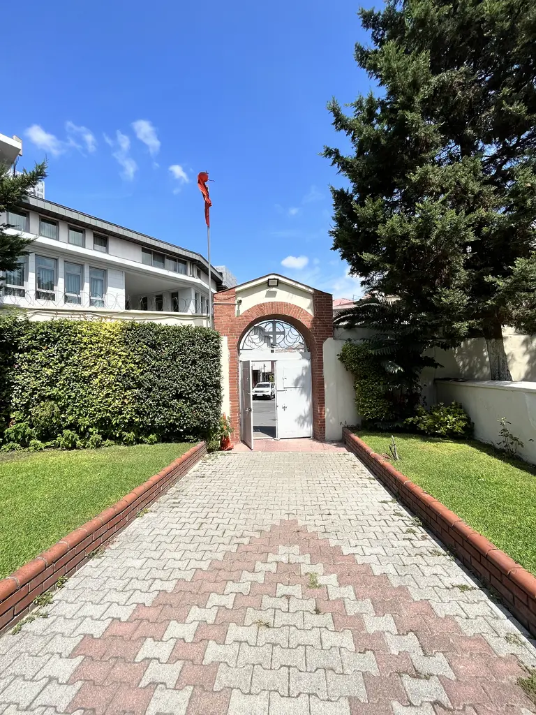 Bahçe yolu; iki yanında budanmış çalı ve çimen, ortada kırmızı desenli taş döşemeli yol ve sonunda tuğla kemerli sokak kapısı.