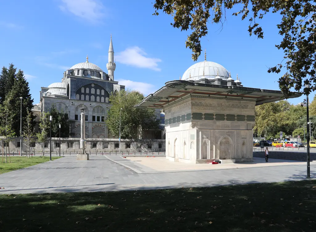 Tophane Meydanı'nda Tophane Çeşmesi; geniş saçaklı kare mermer gövde, yeşil zeminli yazı kuşağı, arkada Kılıç Ali Paşa Camii'nin kubbesi ve minaresi.