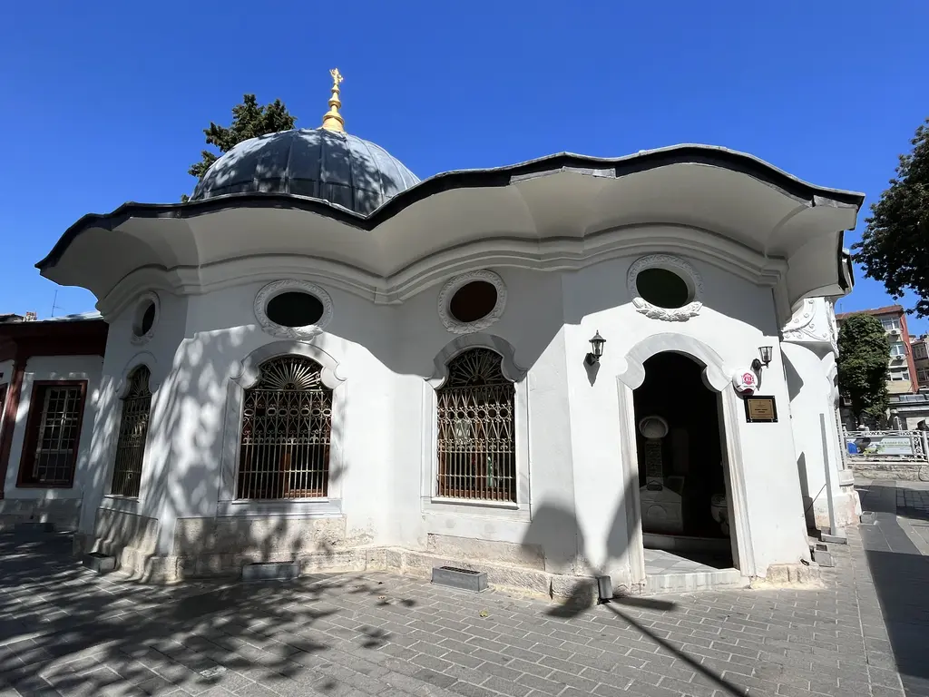 Koca Mustafa Paşa Külliyesi'nde bir türbenin dış cephesi; beyaz sıvalı gövde, barok dalgalı saçak, yuvarlak öküzgözü pencereler, şebekeli kemerli pencereler ve üstte kurşun kubbe.
