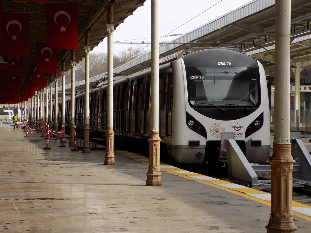 Sirkeci Garı'nın tarihi peronunda beyaz kırmızı banliyö treni; yuvarlak burun, sütunlar arasına asılmış Türk bayrakları, ıslak gri zeminde sarı güvenlik şeridi ve solda ikinci tren.